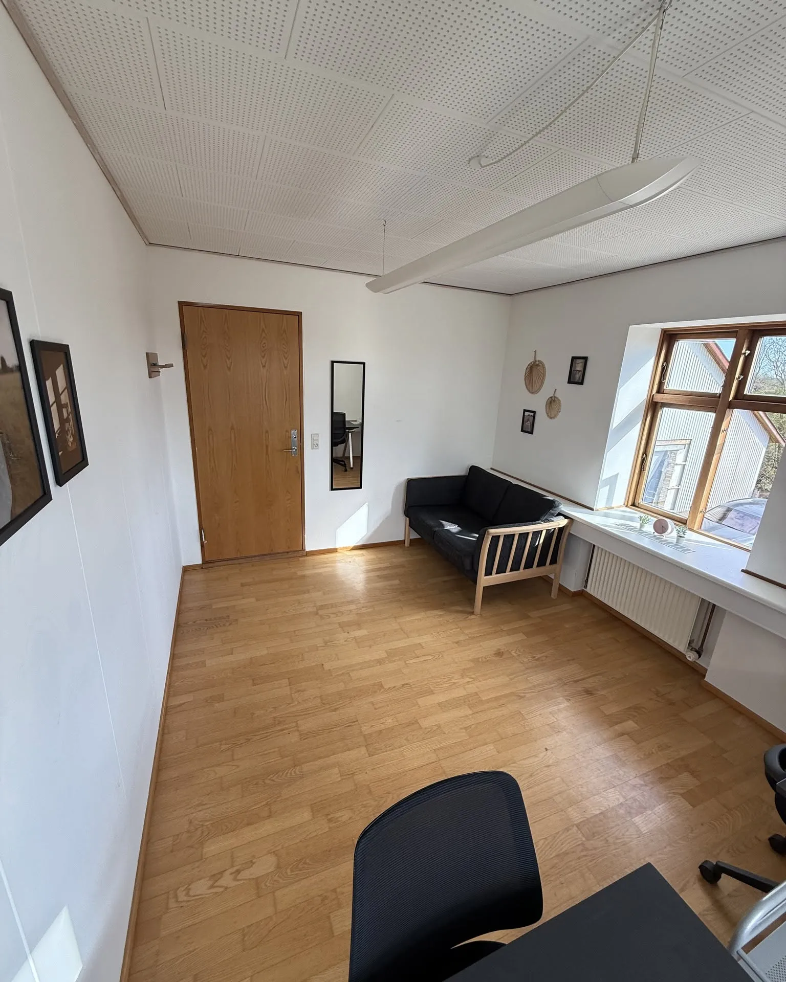 Kontorlokale på Hundtofte 14A med trægulv, akustikloft, sofa og vindue, set fra skrivebordet