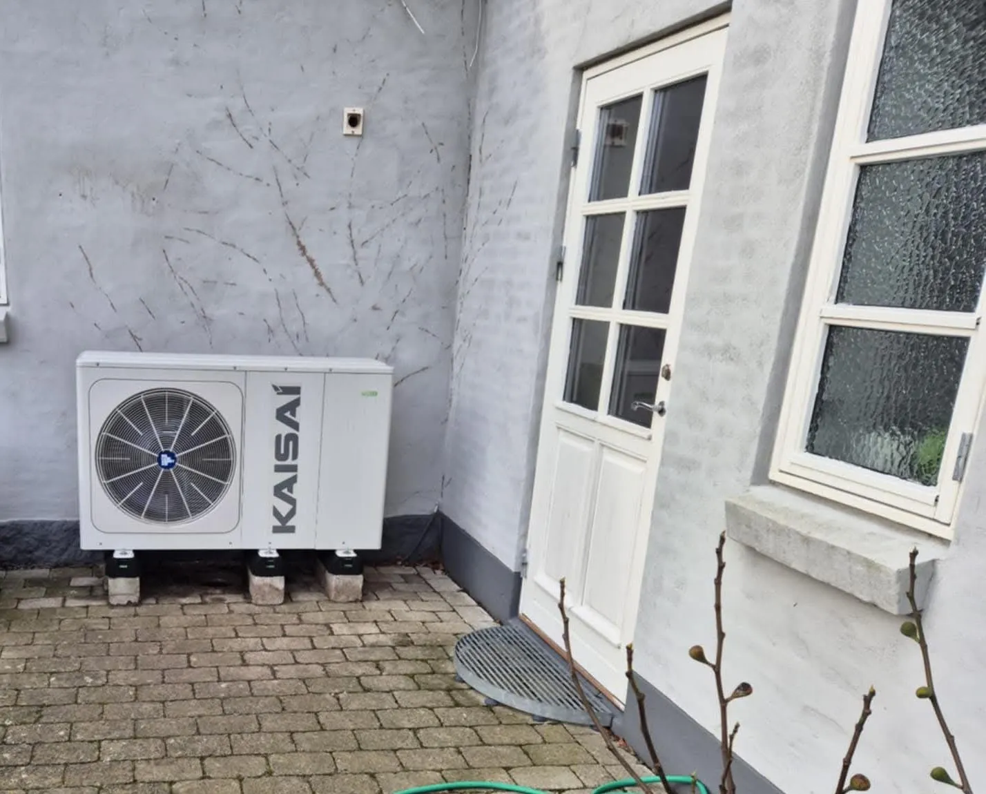 Varmepumpens udedel i en brolagt gårdsplads ved bagdøren