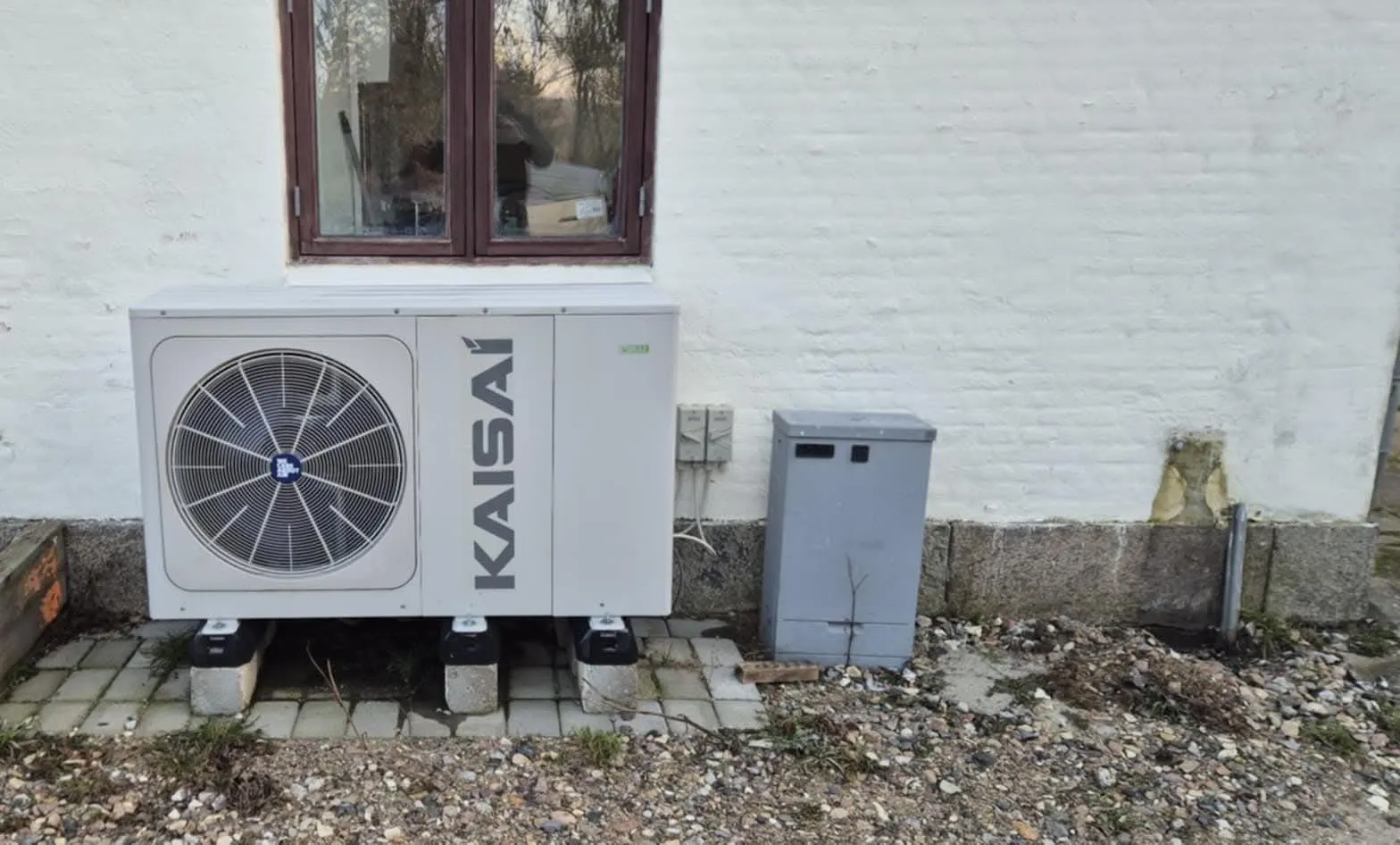 Varmepumpens udedel under et vindue på en hvid kalket mur
